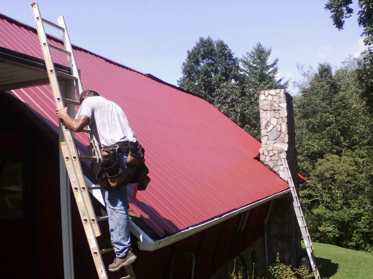 Roofer on a ladder beside a red metal roof and stone chimney — Soffit & Fascia Repair by Roofing Brothers in Montebello